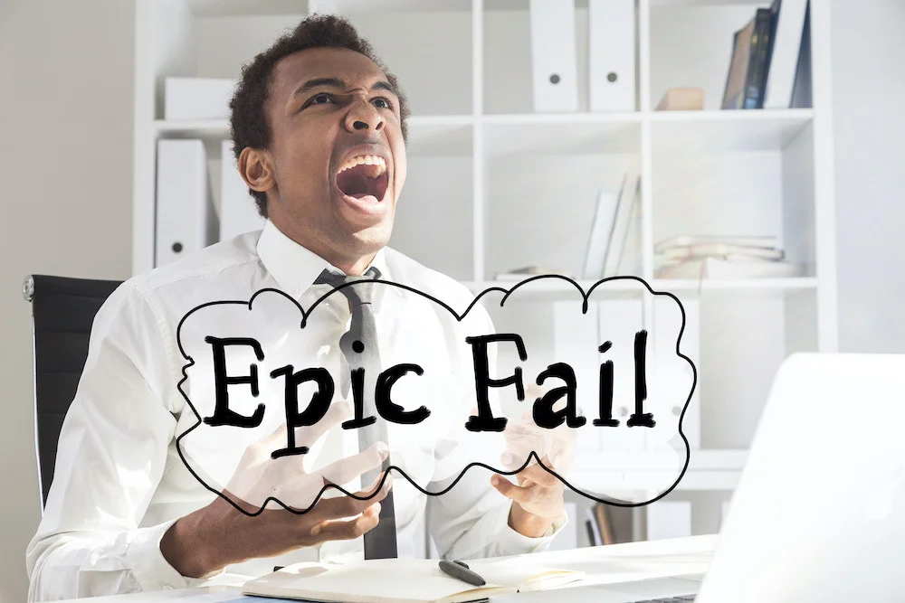 Concept of failure with screaming furious black businessman at office desk with laptop, notepad and 'epic fail' text