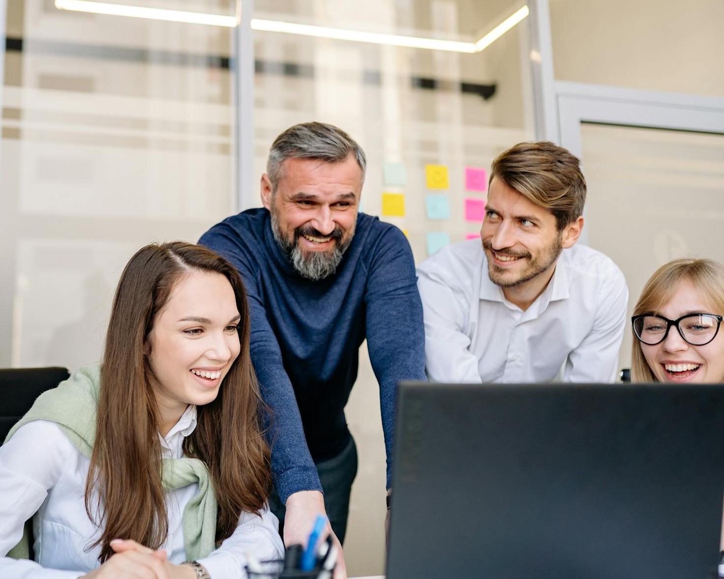 Tým se směje u počítače, manažer stojí za nimi a sleduje výsledky, znázorňuje spolupráci a společnou radost z úspěchu.